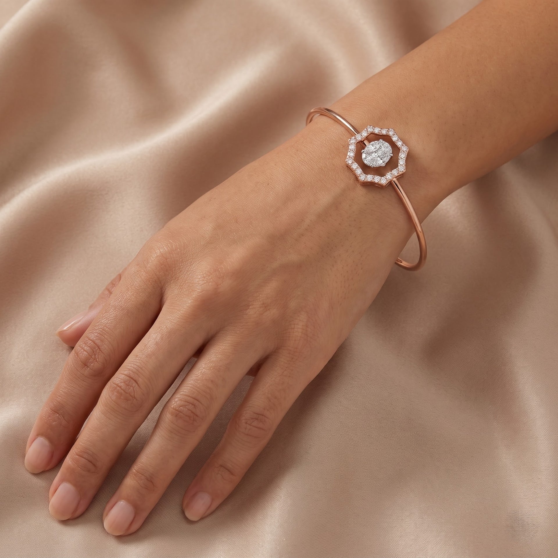 A rose gold cuff bracelet with a central halo of small clear stones and a round center stone, shown worn on a hand against a draped beige satin background.