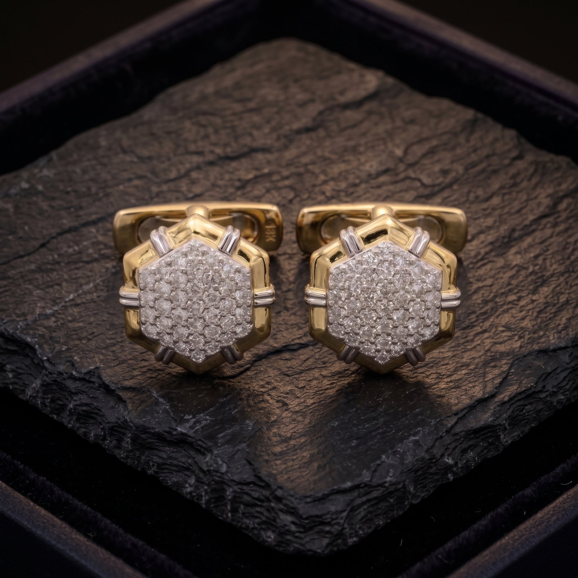A pair of gold-toned cufflinks with pavé-set clear stones, displayed on a dark textured stone slab inside a black presentation box.