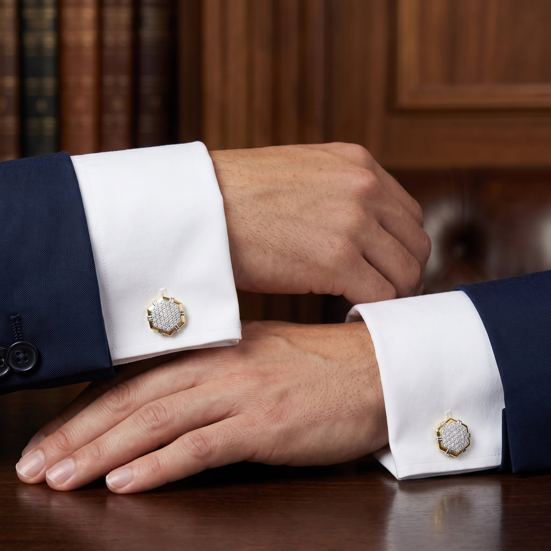 White dress shirt cuffs with gold cufflinks, worn with navy blue suit jackets, resting on a dark wooden table.