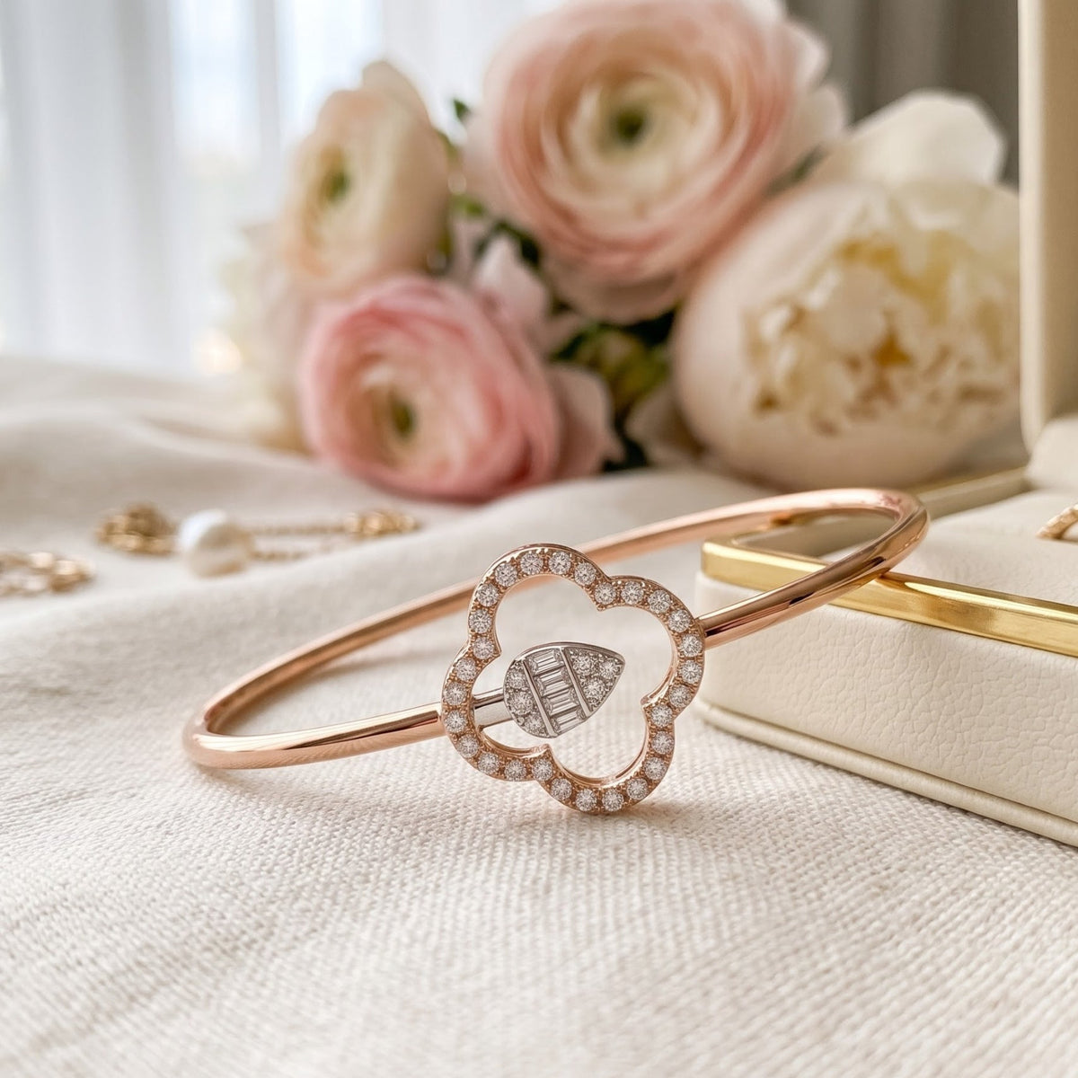 A rose gold bangle bracelet with a heart-shaped pavé frame and a central chevron-patterned diamond, displayed on a textured cream fabric with soft pink flowers and jewelry in the background.