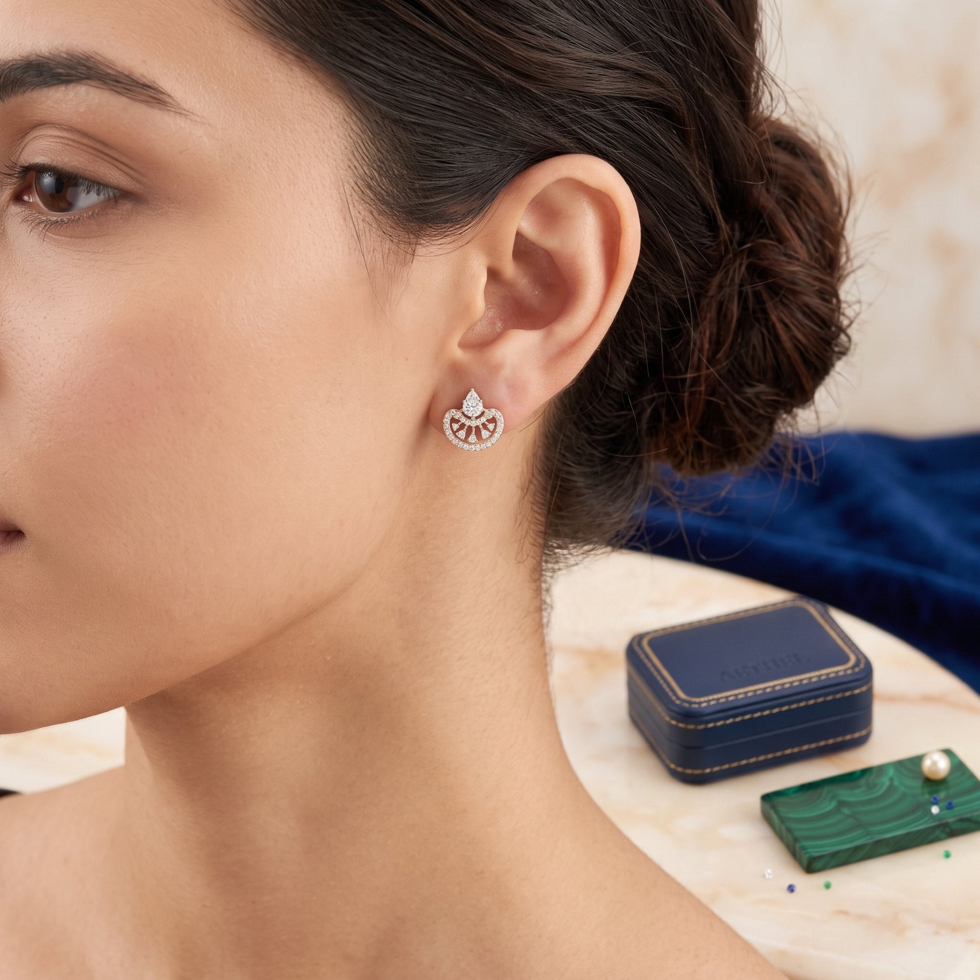 A close-up lifestyle shot of a woman wearing a delicate silver-toned earring with a fan-shaped pavé setting. A small navy blue jewelry box and a green pearl card are visible on a marble table in the background.