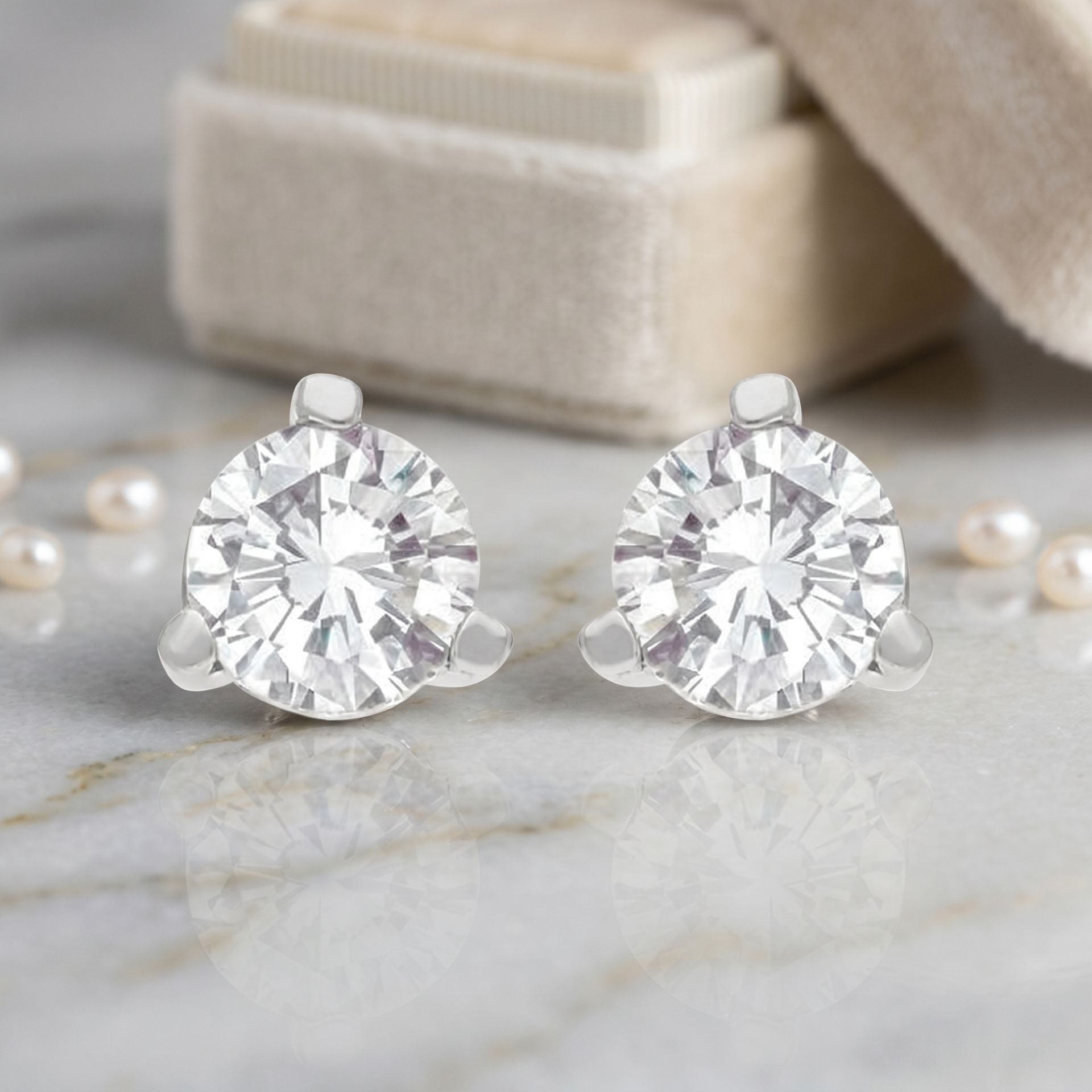 Pair of diamond stud earrings on a marble surface with pearls and a beige box in the background.