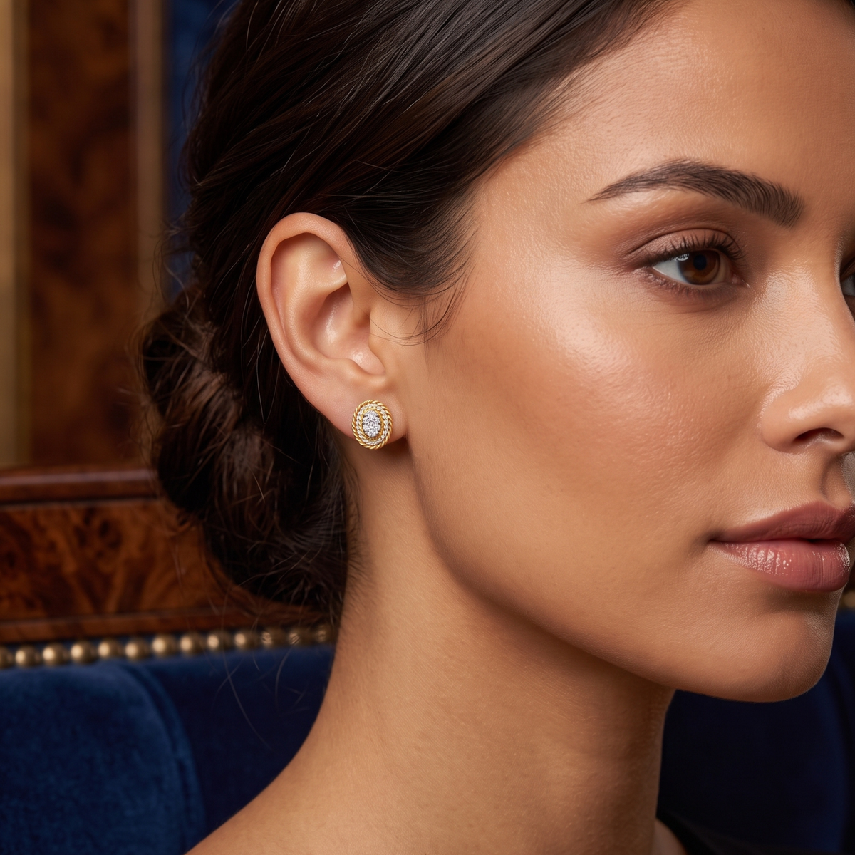 A close-up side profile of a woman wearing a small oval stud earring with a gold-toned border and a central white stone, set against a dark blue velvet and wood background.