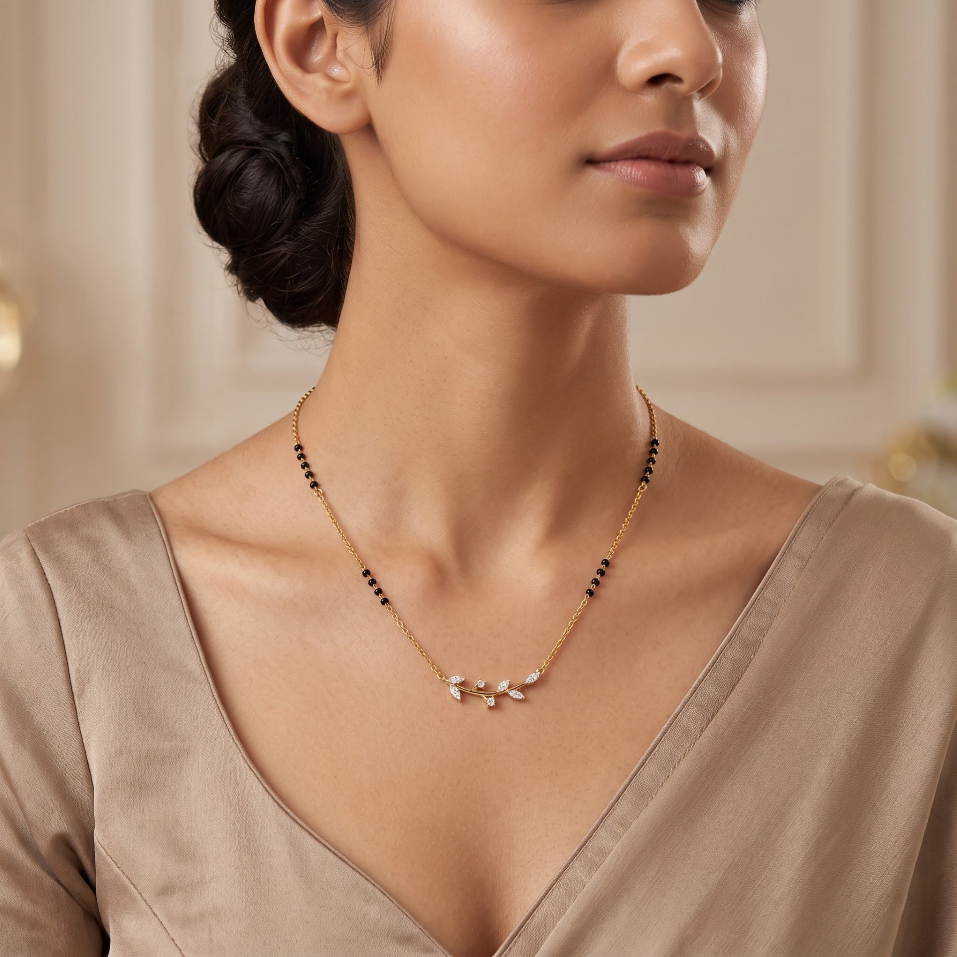 A close-up of a woman wearing a delicate gold necklace with small black beads and a central white floral or leaf-like cluster pendant, paired with a taupe satin top.