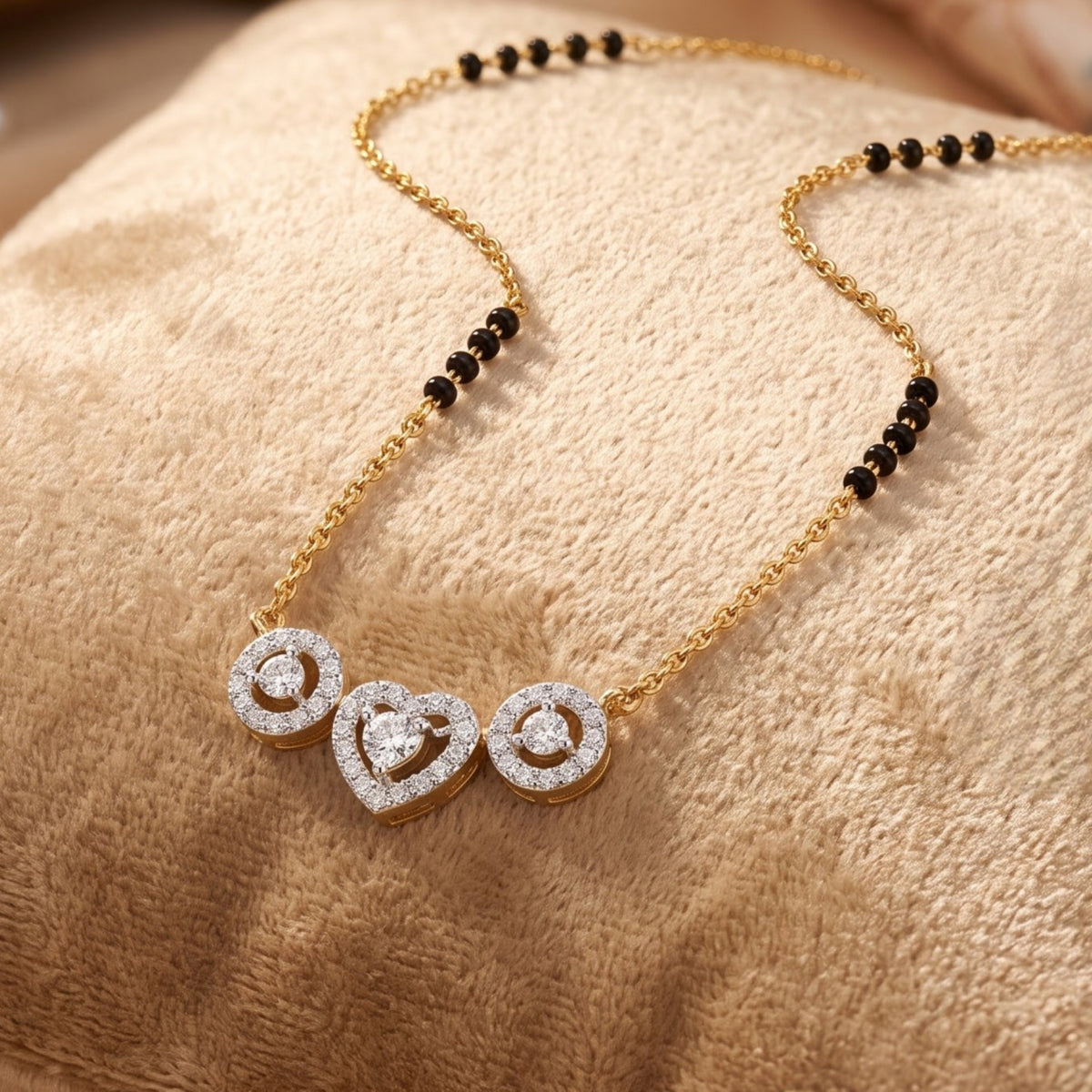 A gold chain necklace with a beaded section of small black beads and a pendant featuring three heart and circle motifs paved with clear stones, displayed on a textured beige fabric surface.