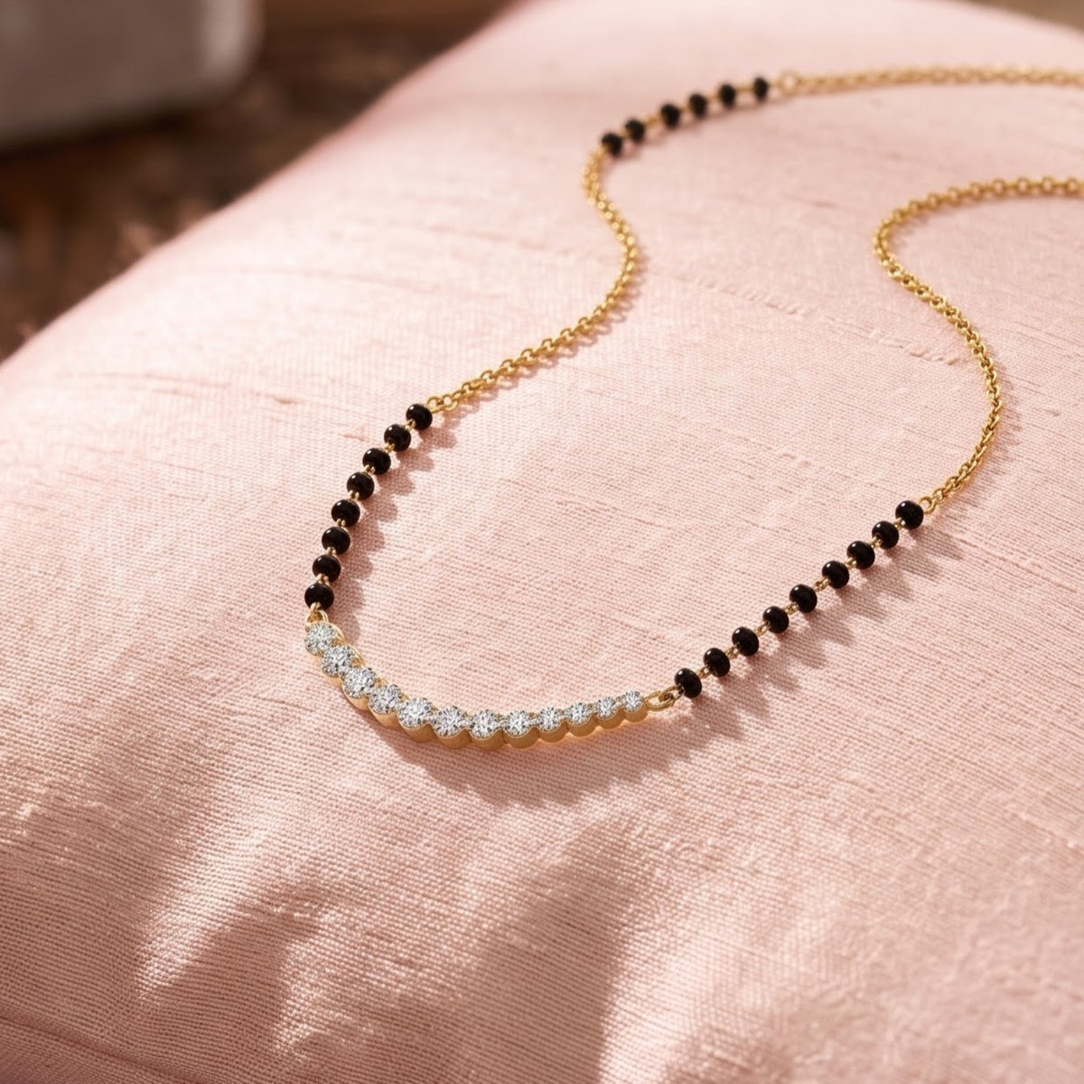 A delicate gold chain necklace with a curved section of small black beads and a section of clear stones, displayed on a soft pink fabric surface.