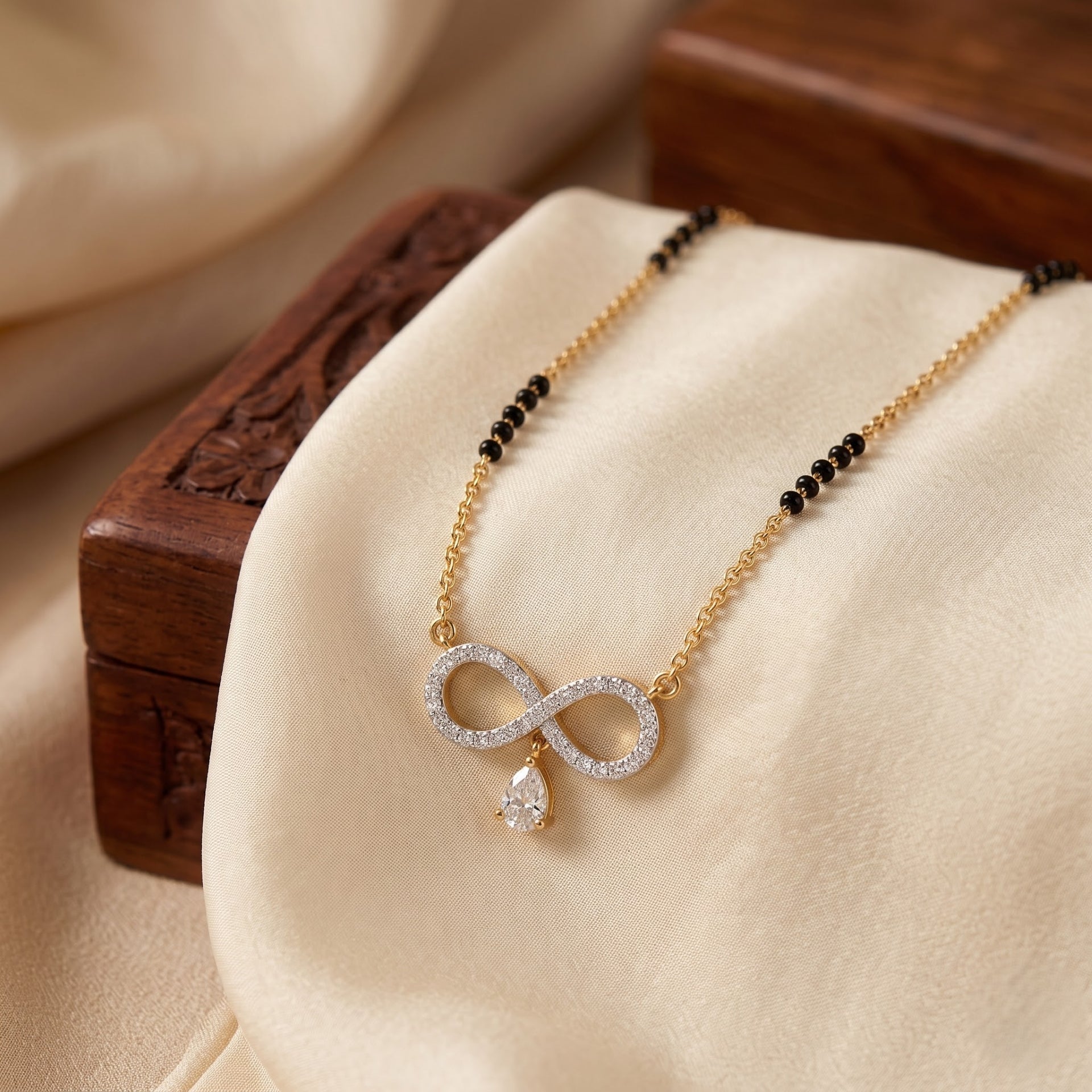 A gold chain necklace with a double-strand black bead section and an infinity-shaped pendant paved with clear stones, featuring a small teardrop crystal drop, displayed on cream satin fabric over carved wooden boxes.