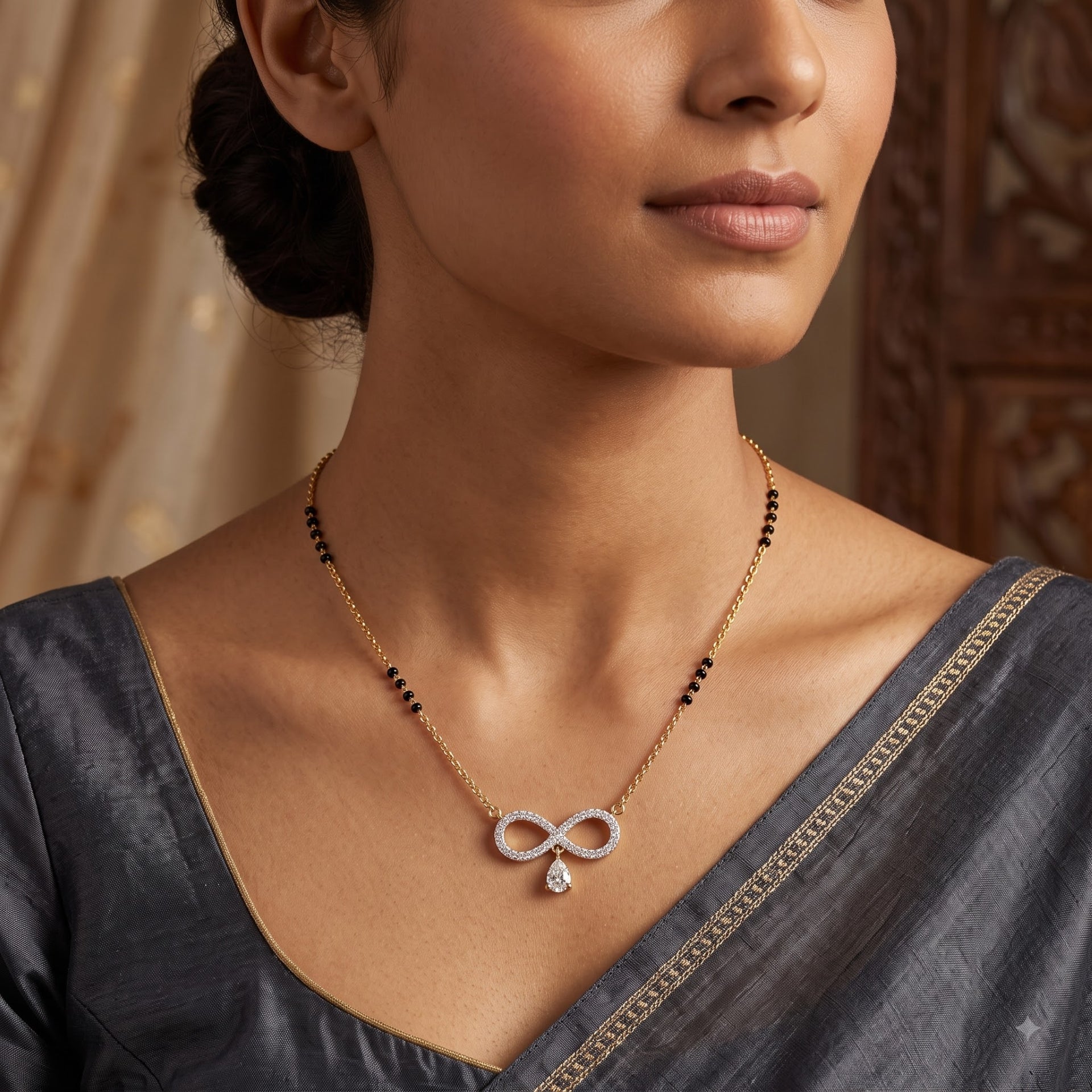 A close-up lifestyle shot of a woman wearing a delicate gold chain necklace with a silver infinity symbol pendant set with small clear stones, and a small teardrop crystal drop. The necklace has black bead accents on the chain.