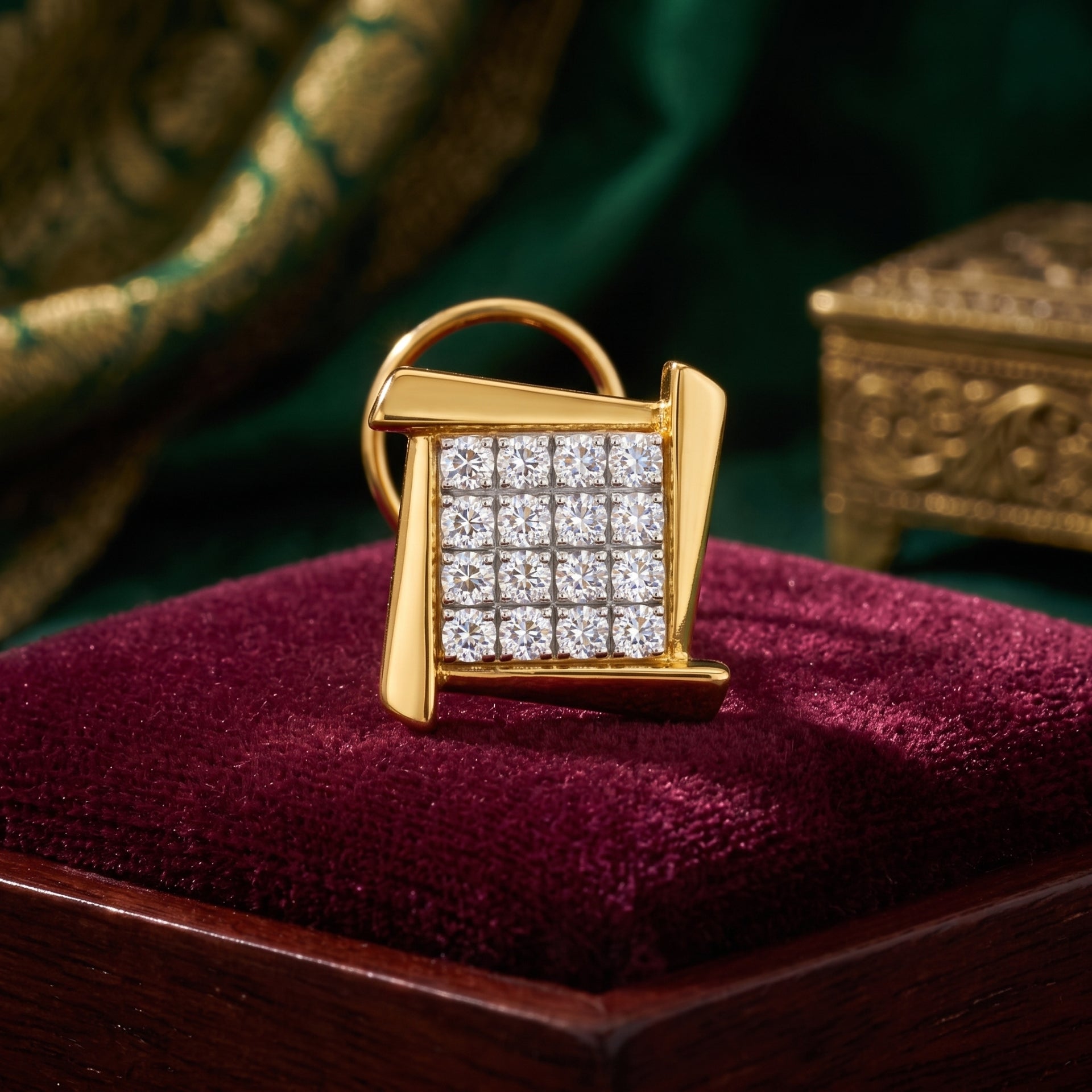 A gold ring with a square face paved with small clear stones, displayed on a deep burgundy velvet jewelry box cushion. The background features dark green draped fabric and a blurred ornate gold box.