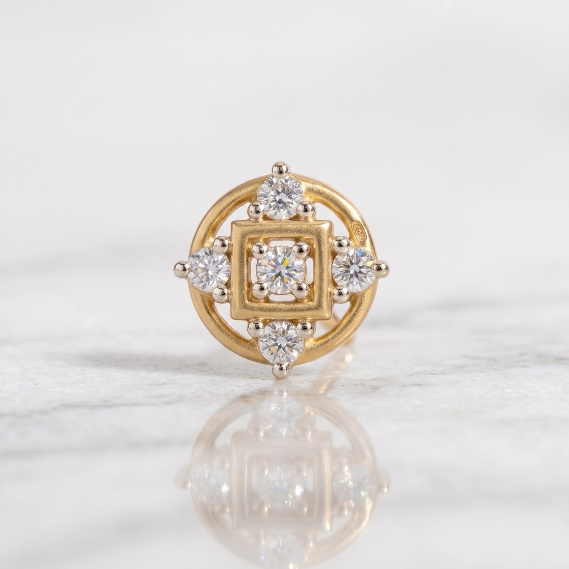 A yellow gold nose ring with a square and circular diamond setting, displayed on a light marble surface with a soft reflection.