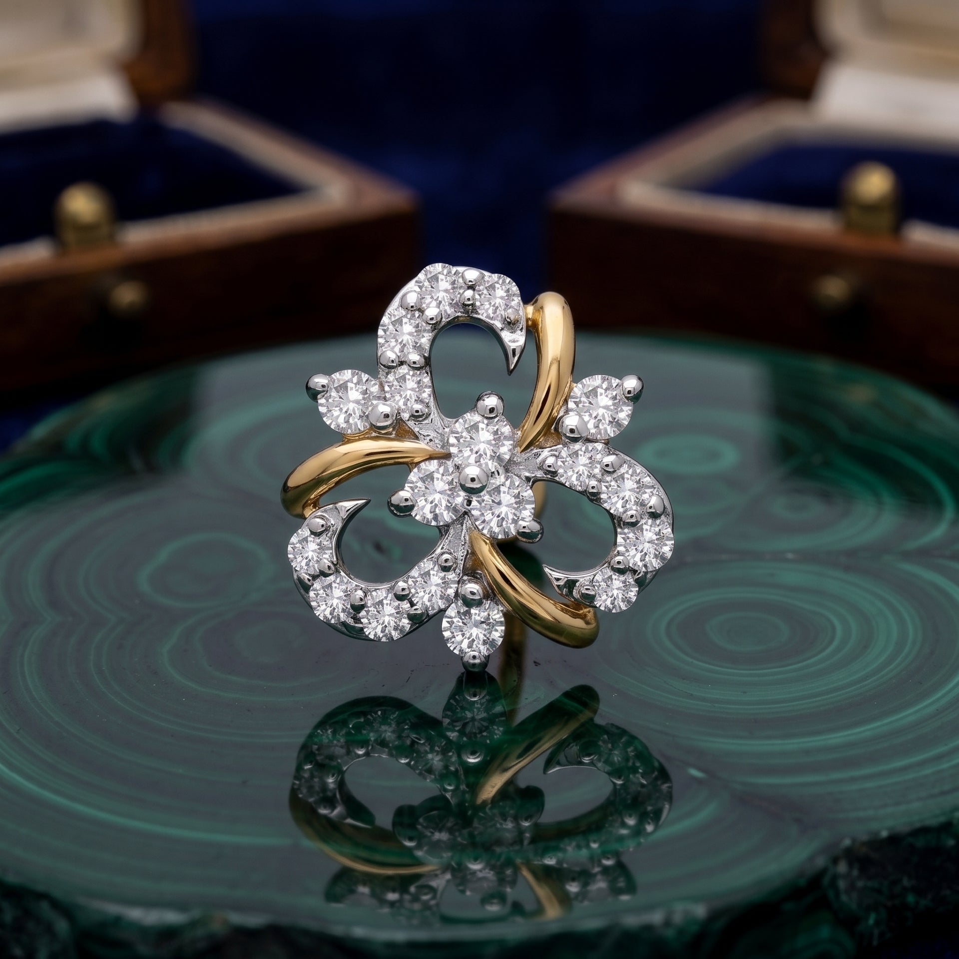A floral-shaped ring with white gold or platinum pavé-set clear stones and curved yellow gold accents, displayed on a green malachite slab with blurred jewelry boxes in the background.