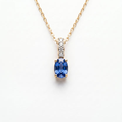 A gold necklace with an oval blue sapphire pendant and a pavé-set diamond accent bar, on a wite background