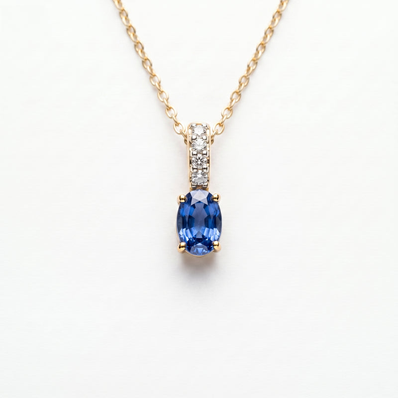 A gold necklace with an oval blue sapphire pendant and a pavé-set diamond accent bar, on a wite background