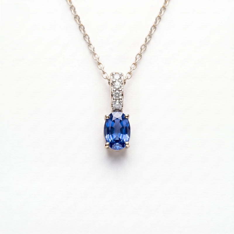 A silver chain necklace with a pendant featuring a marquise-cut blue sapphire gemstone and a small row of clear accent stones above it, set against a plain white background.