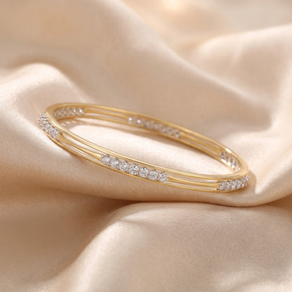 A gold bangle bracelet with rows of small clear stones, displayed on draped beige satin fabric.