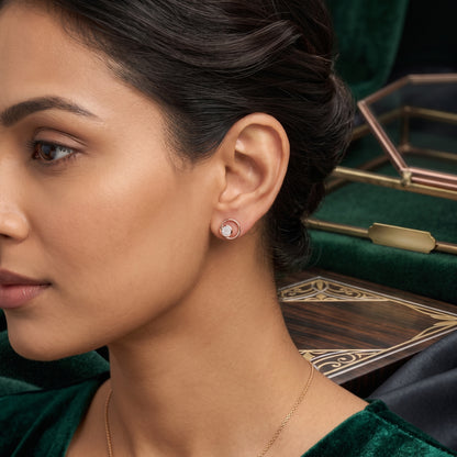 A close-up of a woman wearing a small round stud earring with a clear gemstone in a gold-toned setting, paired with a delicate gold necklace and a dark green velvet top. A matching green velvet jewelry box is visible in the background.