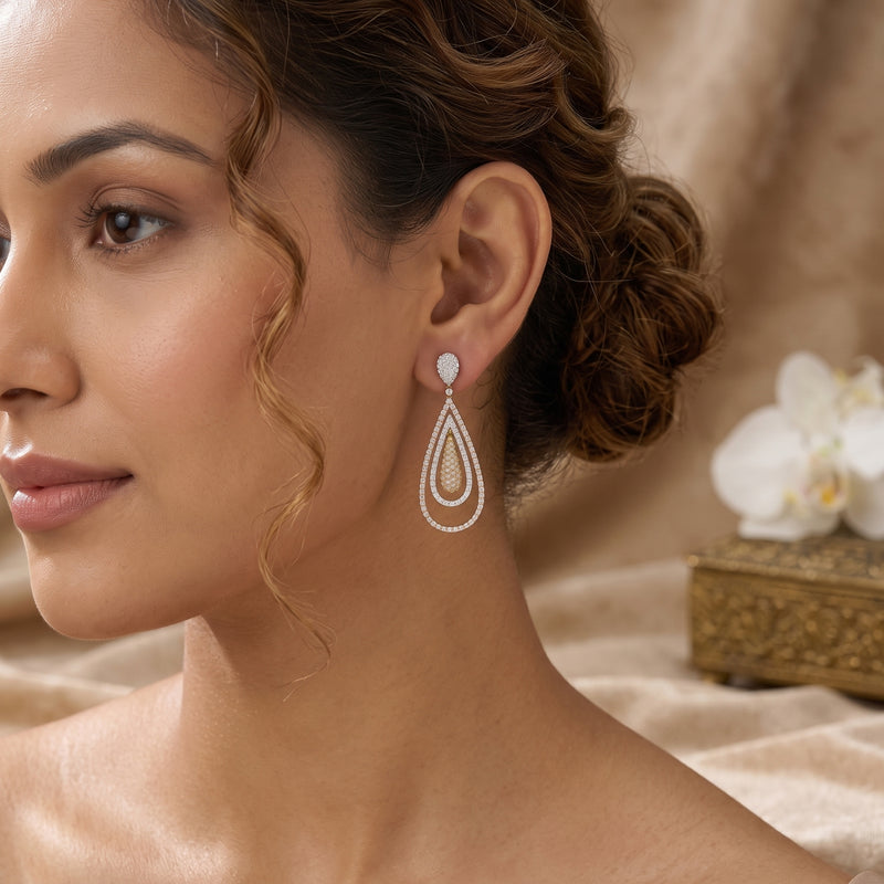 A woman wearing a teardrop-shaped drop earring with a white stone and silver-toned layered detailing, shown in a close-up portrait against a soft beige background with a white orchid flower.