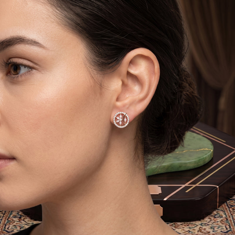 A close-up side profile of a woman wearing a small round stud earring with a silver-toned circular setting and a central pink gemstone surrounded by clear stones.