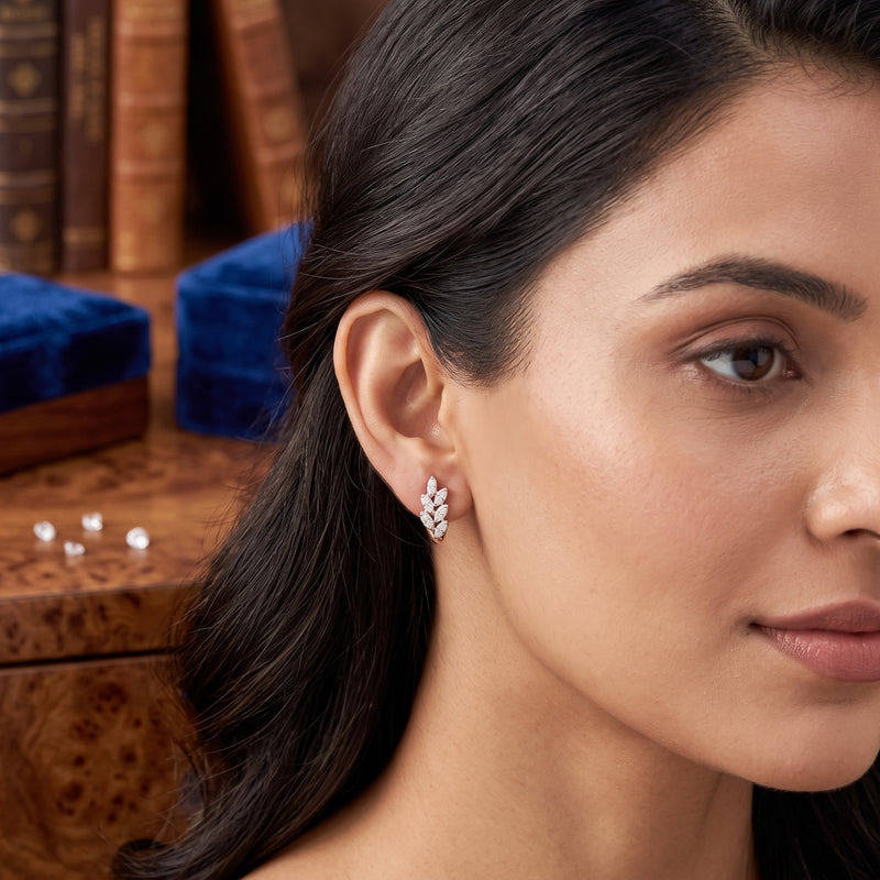 A close-up of a woman wearing a delicate silver-toned leaf-shaped stud earring with small clear stones, set against a warm, blurred background of books and velvet boxes.