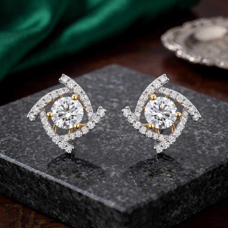 A pair of gold and silver spiral stud earrings with clear round-cut center stones and small clear accent stones, displayed on a dark stone block with a green satin fabric in the background.