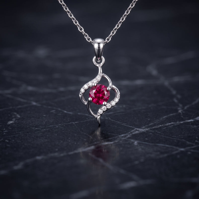 A silver pendant necklace featuring a central round red gemstone surrounded by small clear stones in an open, flowing design, hanging from a silver chain on a dark marble surface.