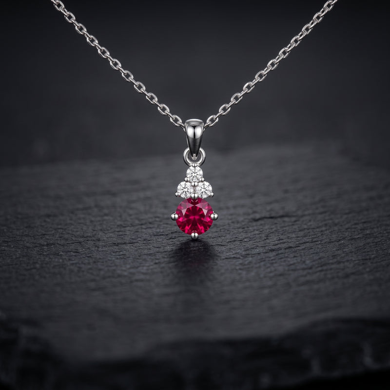 A silver pendant necklace featuring a round red gemstone and small clear accent stones, displayed on a dark textured surface.