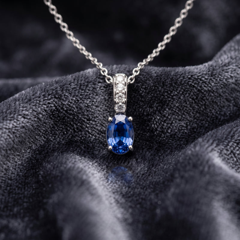A silver necklace with an oval-cut blue sapphire pendant and a small halo of clear stones, displayed on a dark textured velvet fabric.