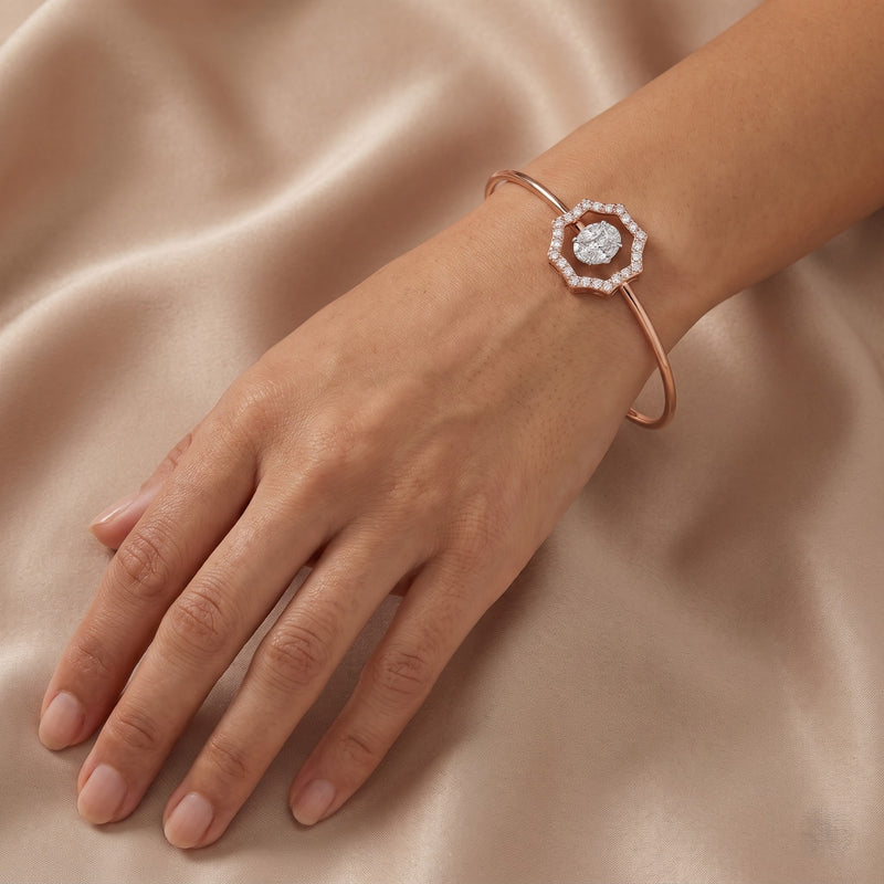 A rose gold cuff bracelet with a central halo of small clear stones and a round center stone, shown worn on a hand against a draped beige satin background.