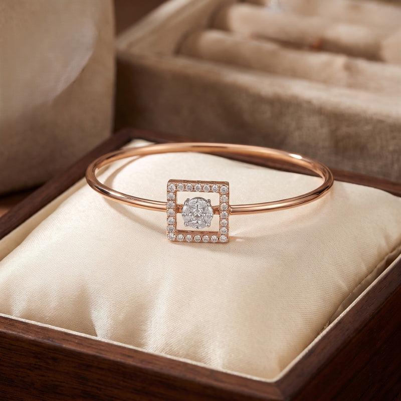 A rose gold bangle bracelet with a square diamond centerpiece, displayed on a cream satin cushion inside a wooden jewelry box.
