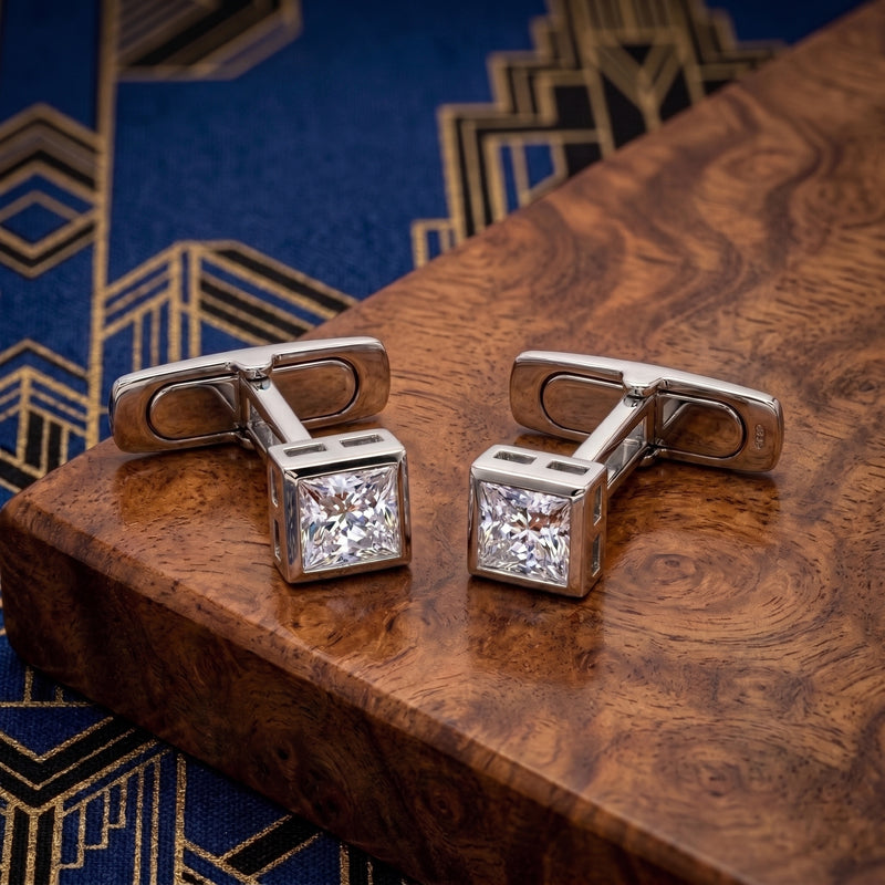 A pair of silver-toned cufflinks with square clear gemstones, displayed on a polished wooden block over a blue patterned fabric background.