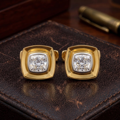 A pair of gold square stud earrings with cushion-cut clear gemstones, displayed on a textured dark brown leather surface.