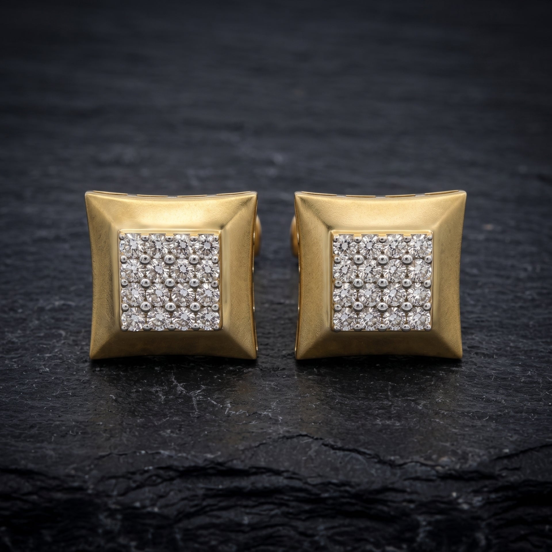 A pair of gold square stud earrings with a central square cluster of clear pavé gemstones, displayed on a dark textured slate surface.