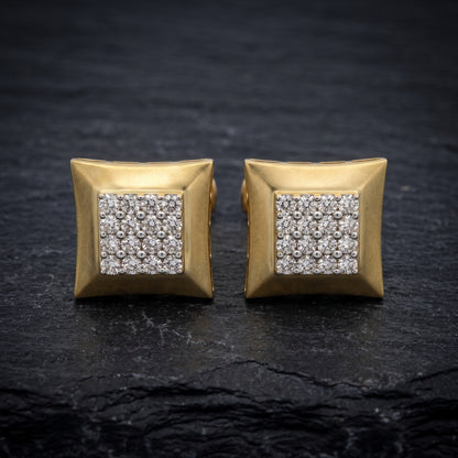 A pair of gold square stud earrings with a central square cluster of clear pavé gemstones, displayed on a dark textured slate surface.