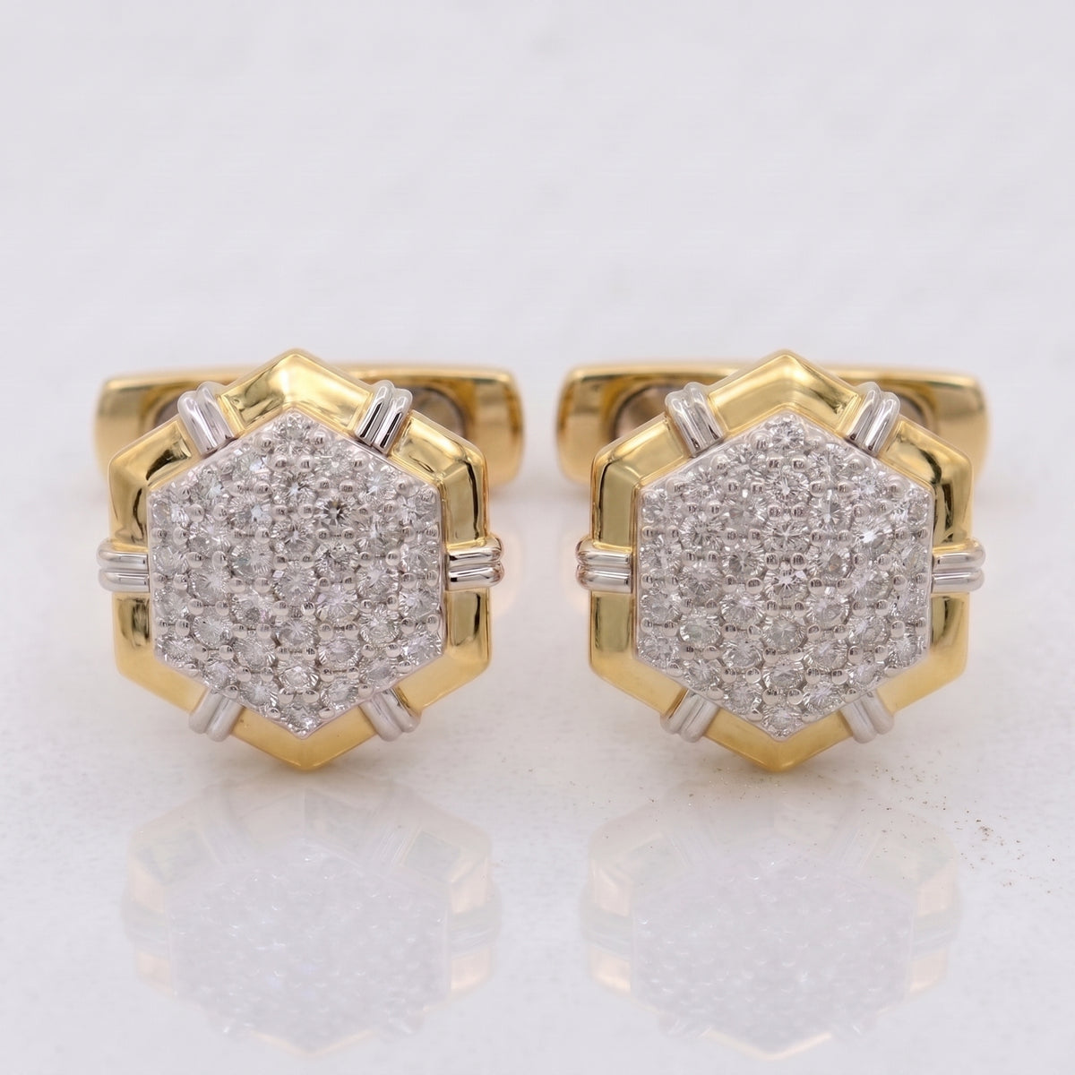 A pair of gold hexagonal stud earrings with pavé-set clear gemstones and small silver-toned side accents, shown on a light reflective surface.