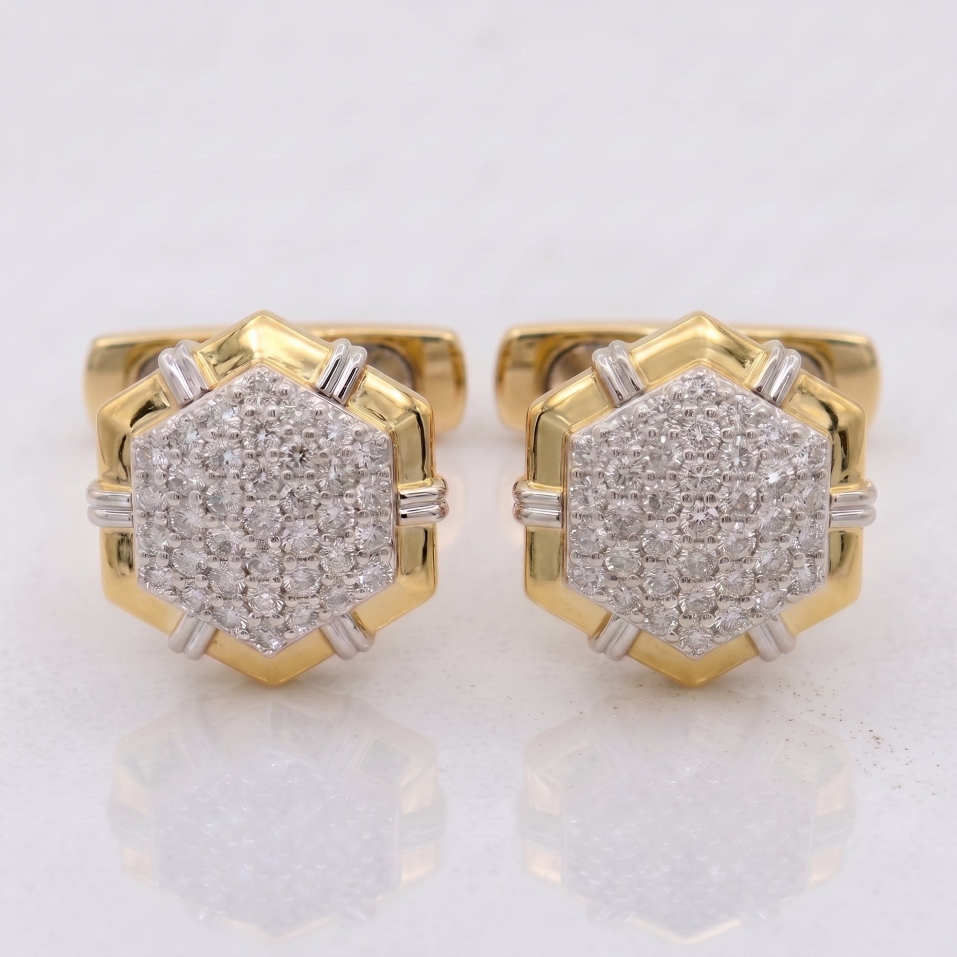 A pair of gold hexagonal stud earrings with pavé-set clear gemstones and small silver-toned side accents, shown on a light reflective surface.