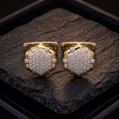 A pair of gold-toned cufflinks with pavé-set clear stones, displayed on a dark textured stone slab inside a black presentation box.