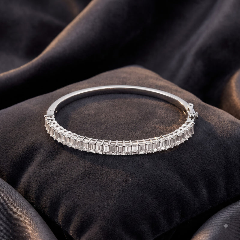 A silver-toned bangle bracelet with a continuous row of baguette-cut clear stones, displayed on a dark brown velvet cushion.