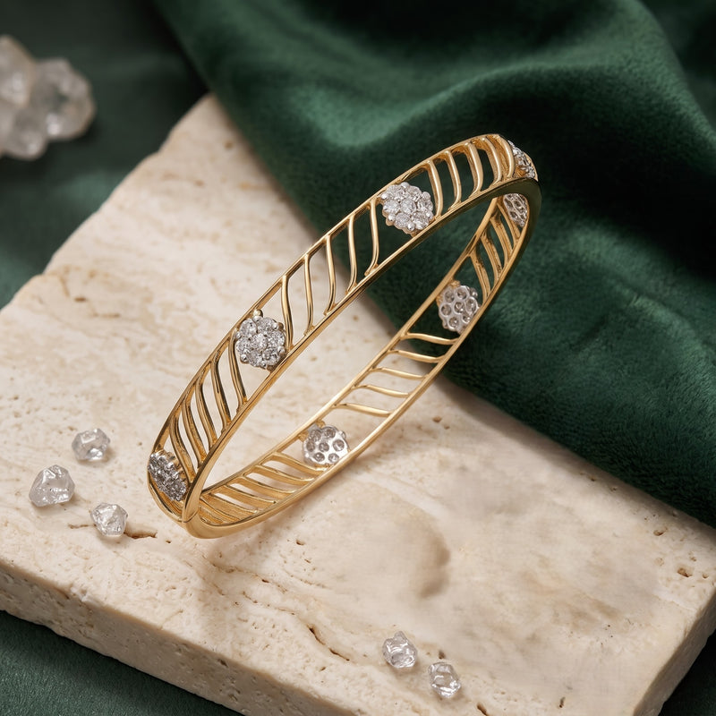 A gold bangle bracelet with a leaf-like openwork pattern and clear crystal accents, displayed on a beige stone slab with a dark green fabric background.