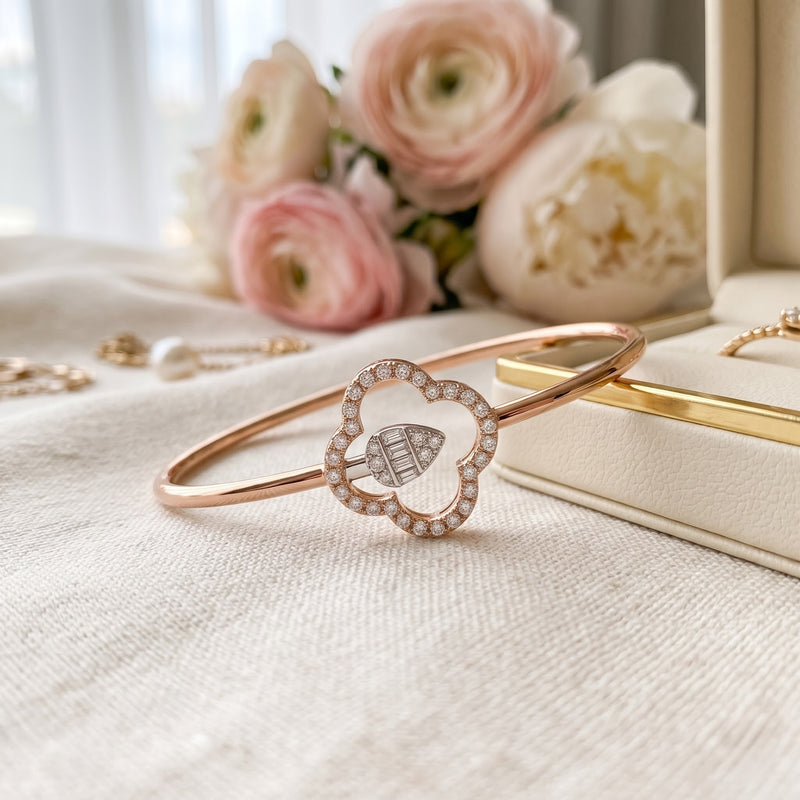 A rose gold bangle bracelet with a heart-shaped pavé frame and a central chevron-patterned diamond, displayed on a textured cream fabric with soft pink flowers and jewelry in the background.