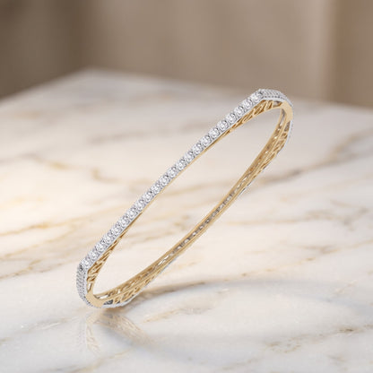 A slim oval bangle bracelet featuring a row of small clear stones on one side and a textured yellow gold finish on the inside, displayed on a light marble surface.