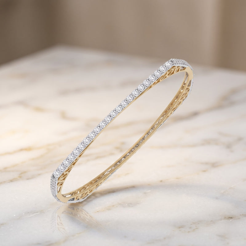A slim oval bangle bracelet featuring a row of small clear stones on one side and a textured yellow gold finish on the inside, displayed on a light marble surface.