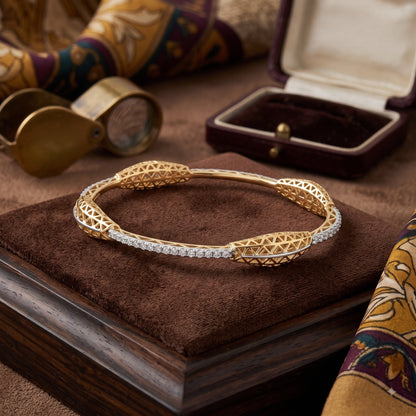 A gold filigree bangle bracelet with a row of clear stones on one side, displayed on a brown velvet jewelry box. The background features a dark velvet jewelry box, a brass loupe, and patterned fabric.