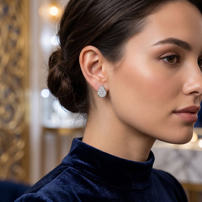 A close-up side profile of a woman wearing a small teardrop-shaped diamond stud earring and a dark navy velvet turtleneck, set against a softly blurred ornate interior background.