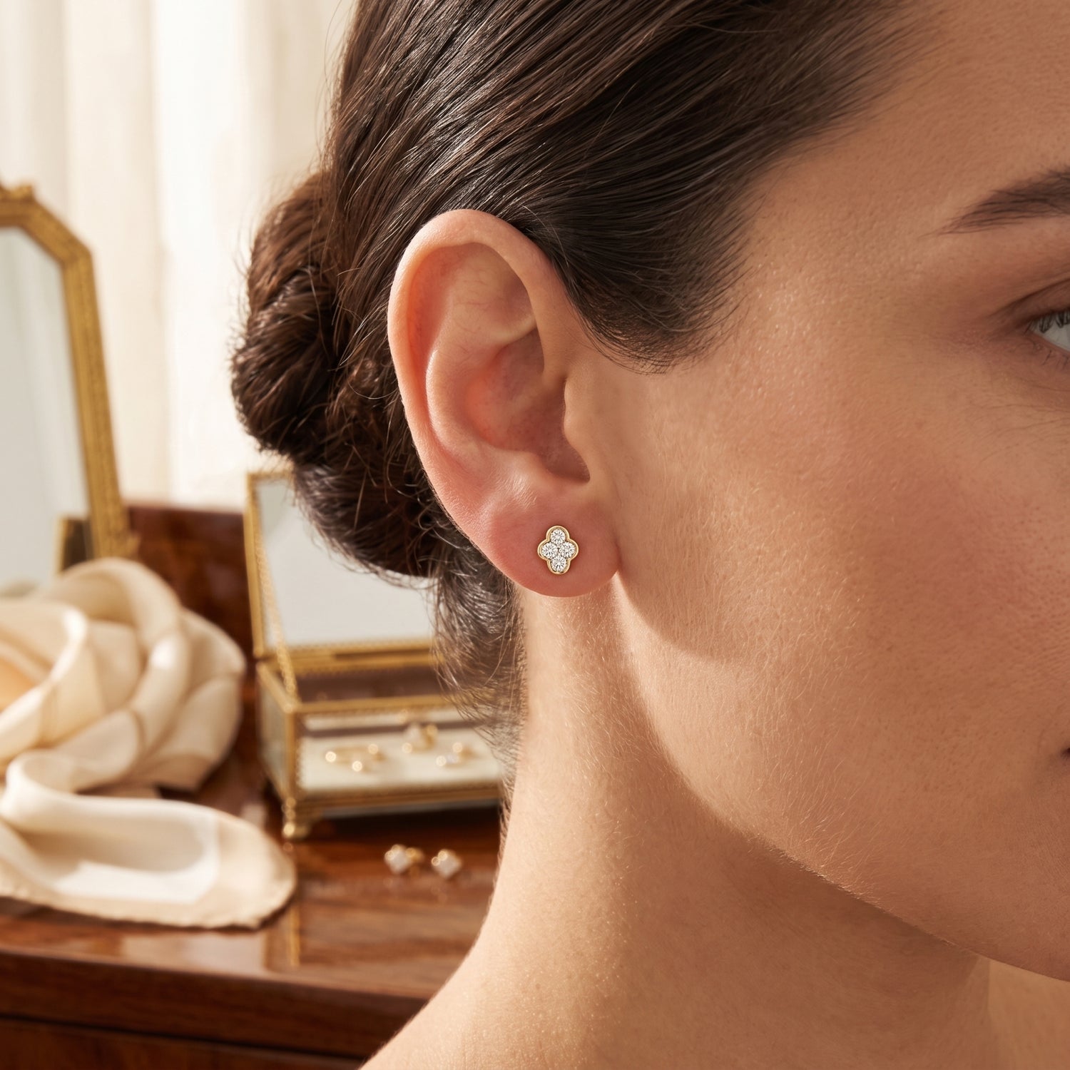 A close-up lifestyle shot of a woman wearing a small four-petal flower stud earring in a warm, elegant room setting.