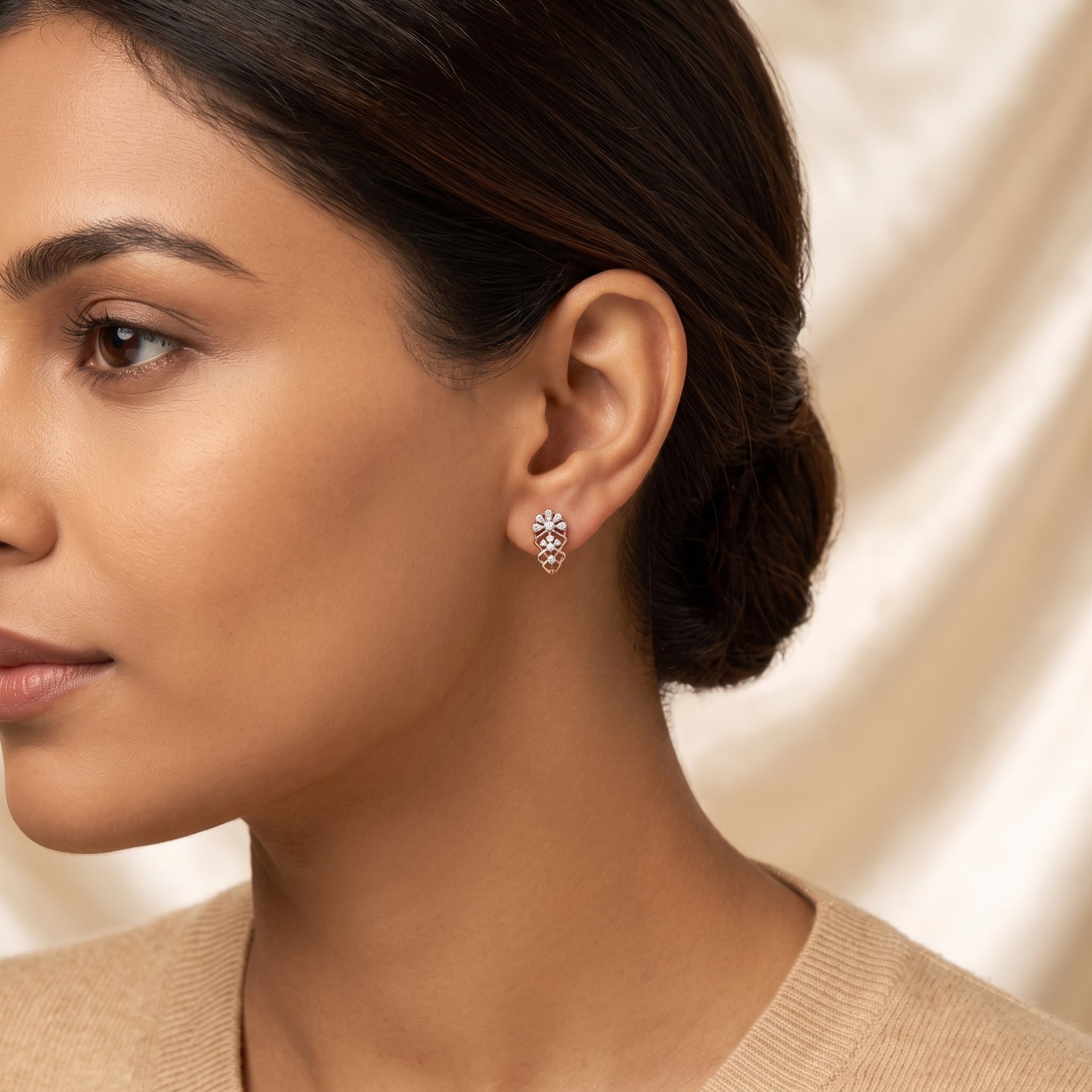 A close-up side profile of a woman wearing a small floral stud earring with clear gemstones in a rose gold setting, against a soft cream satin background.