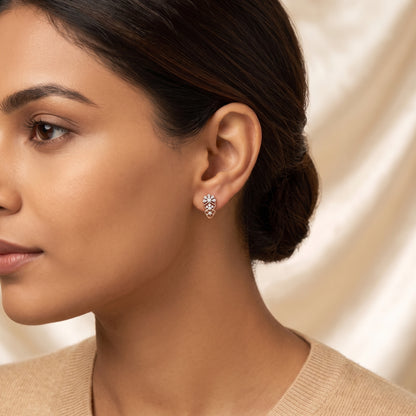 A close-up side profile of a woman wearing a small floral stud earring with clear gemstones in a rose gold setting, against a soft cream satin background.