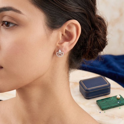 A close-up lifestyle shot of a woman wearing a delicate silver-toned earring with a fan-shaped pavé setting. A small navy blue jewelry box and a green pearl card are visible on a marble table in the background.