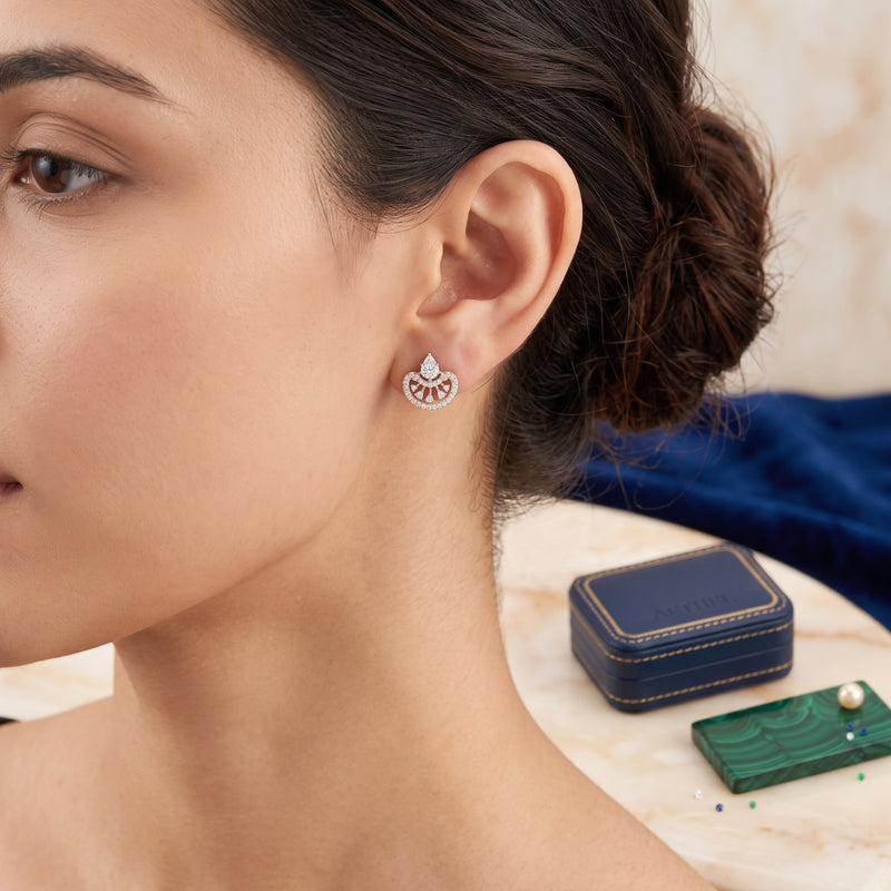 A close-up lifestyle shot of a woman wearing a delicate silver-toned earring with a fan-shaped pavé setting. A small navy blue jewelry box and a green pearl card are visible on a marble table in the background.