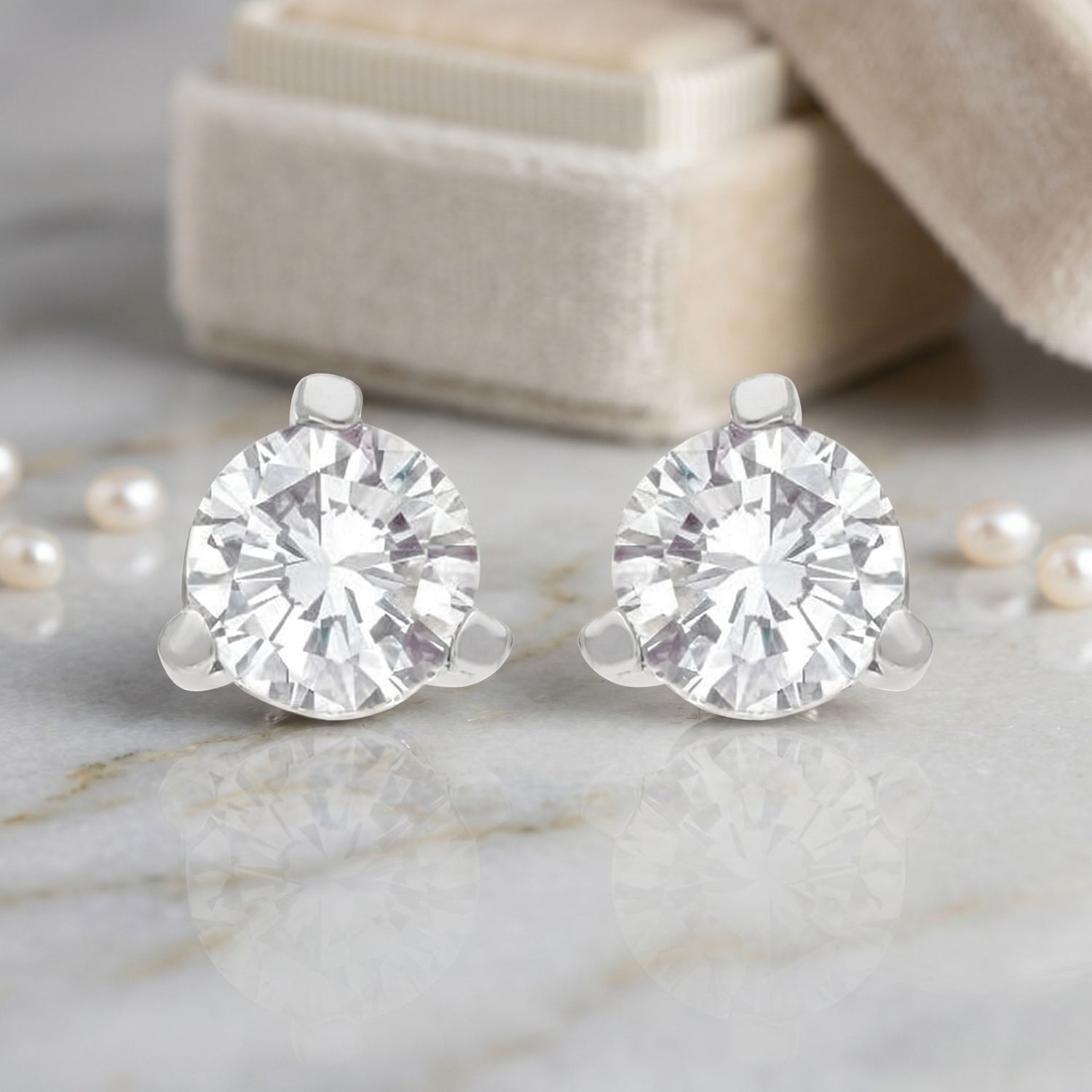Pair of diamond stud earrings on a marble surface with pearls and a beige box in the background.