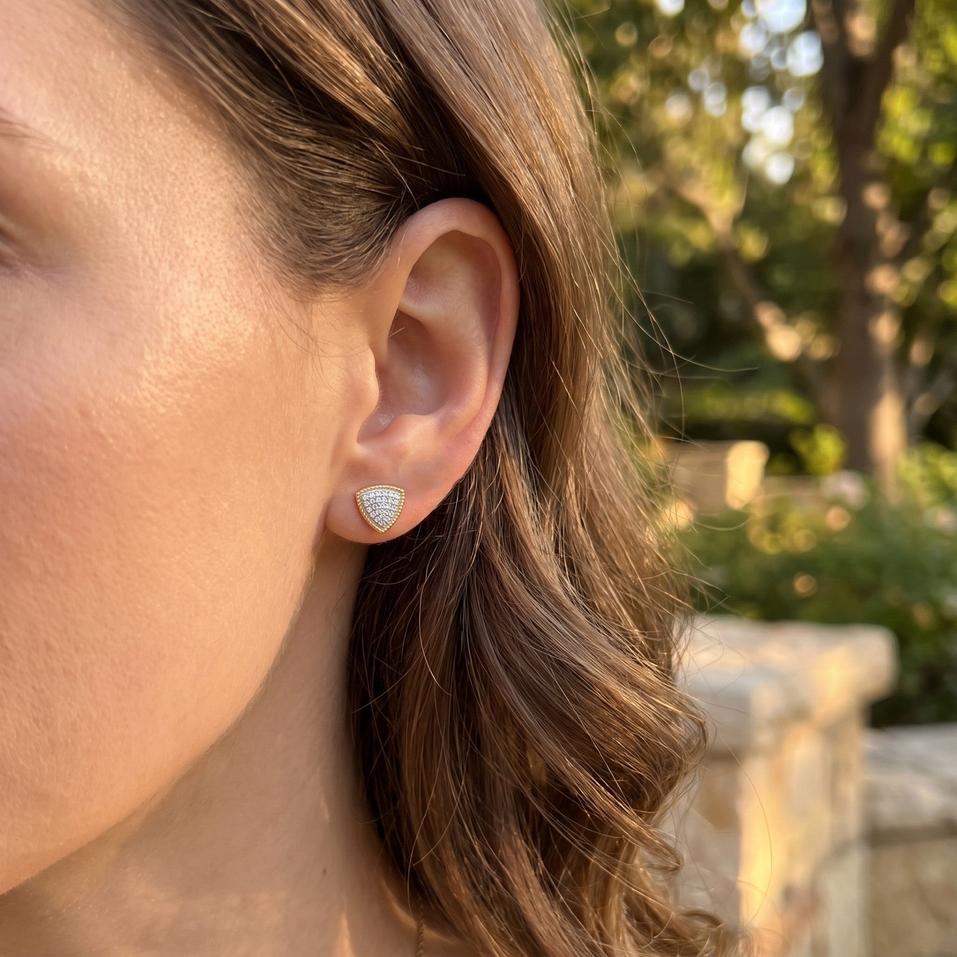 A close-up side profile of a woman wearing a small triangular stud earring with a pavé diamond setting, photographed outdoors with a blurred garden background.